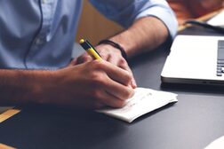 Ein Mann macht sich handschriftliche Notizen, neben ihm auf dem Tisch liegt ein Laptop
